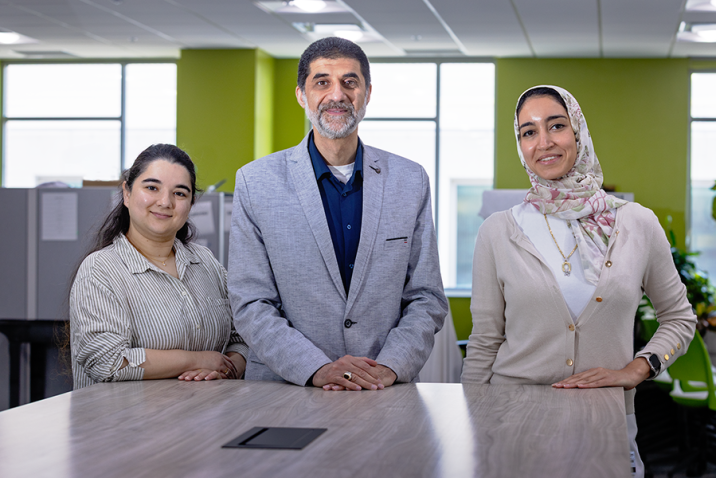 A male scientist and two female scientists stand at the end of a long table in a large, modern office space.