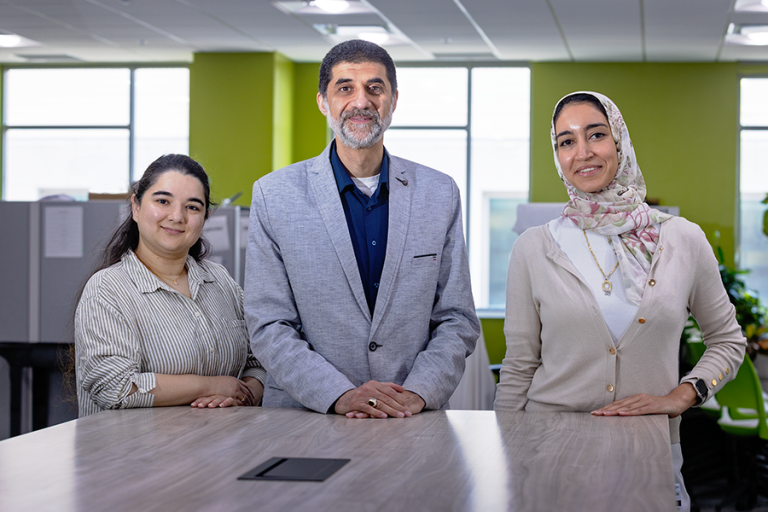 A male scientist and two female scientists stand at the end of a long table in a large, modern office space.