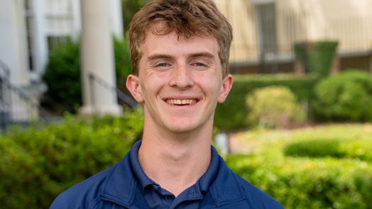 A college-aged man smiling for a photo while standing outside.