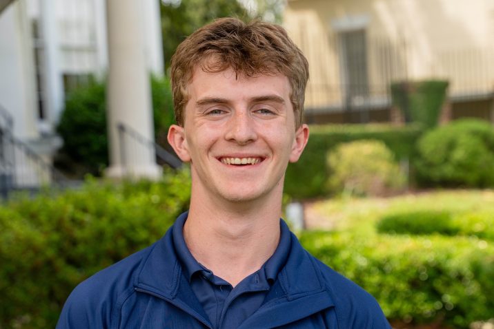 A college-aged man smiling for a photo while standing outside.