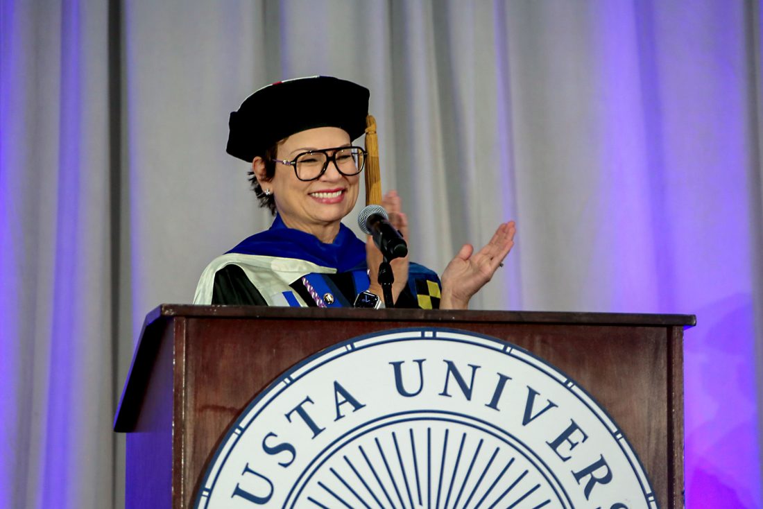 A woman in doctoral regalia claps and smiles while standing behind a podium