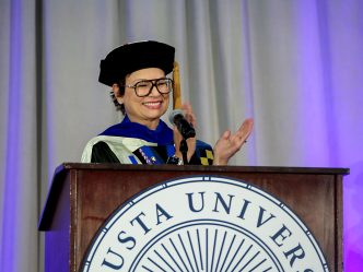 A woman in doctoral regalia claps and smiles while standing behind a podium