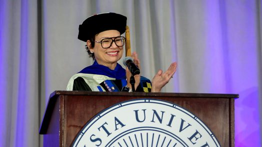 A woman in doctoral regalia claps and smiles while standing behind a podium