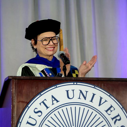 A woman in doctoral regalia claps and smiles while standing behind a podium