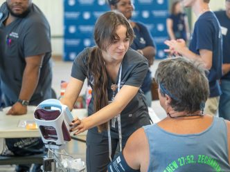 A student in scrubs operates a blood pressure machine while speaking with a participant. They are surrounded by other workers and volunteers.