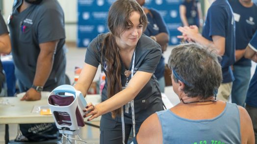 A student in scrubs operates a blood pressure machine while speaking with a participant. They are surrounded by other workers and volunteers.