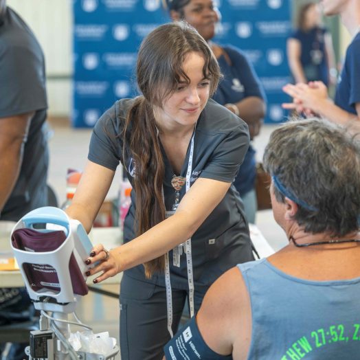 A student in scrubs operates a blood pressure machine while speaking with a participant. They are surrounded by other workers and volunteers.