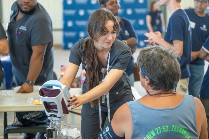 A student in scrubs operates a blood pressure machine while speaking with a participant. They are surrounded by other workers and volunteers.
