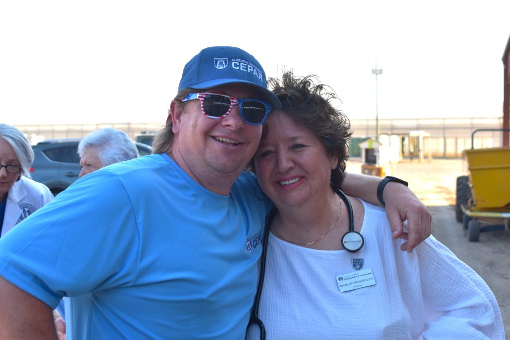 A man and woman pose together and smile at the camera, they are outside on a dirt path.