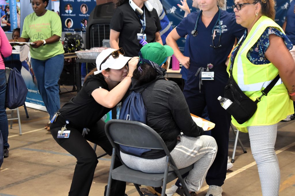 A student inspects the ears of a health fair participant, they are accompanied by an interpreter in a neon vest.