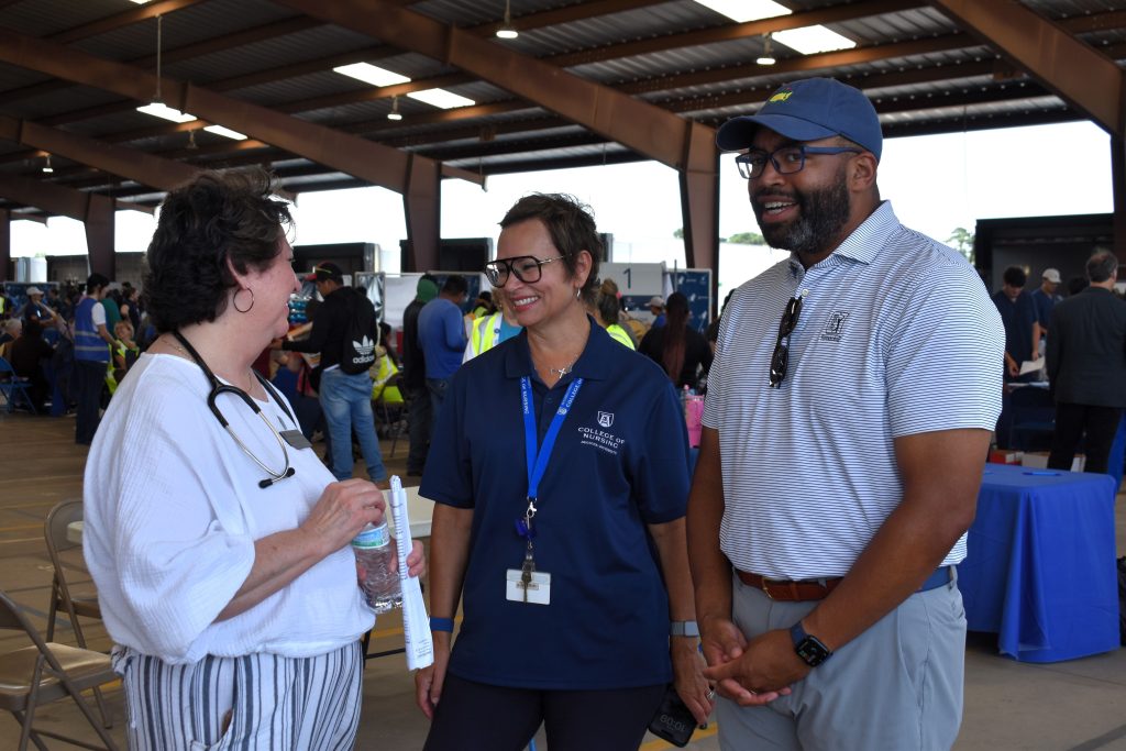 Three people stand together, talking and smiling as a health fair occurs behind them.