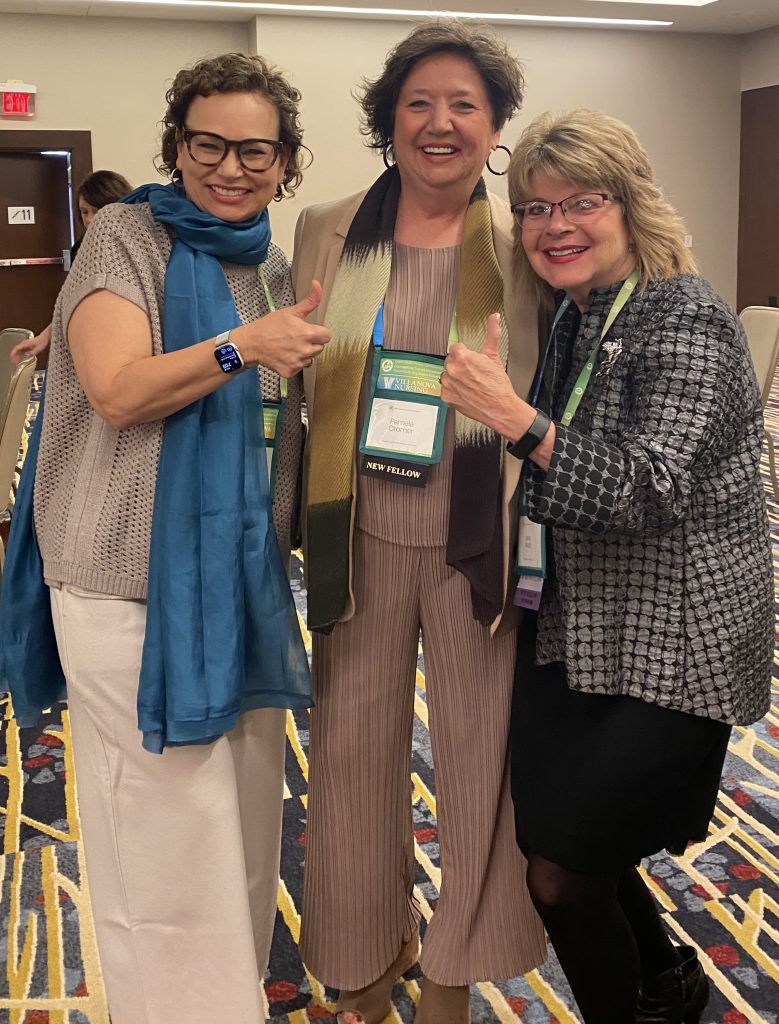 Three women stand together and smile at the camera, the two on the sides are pointing at the one in the middle