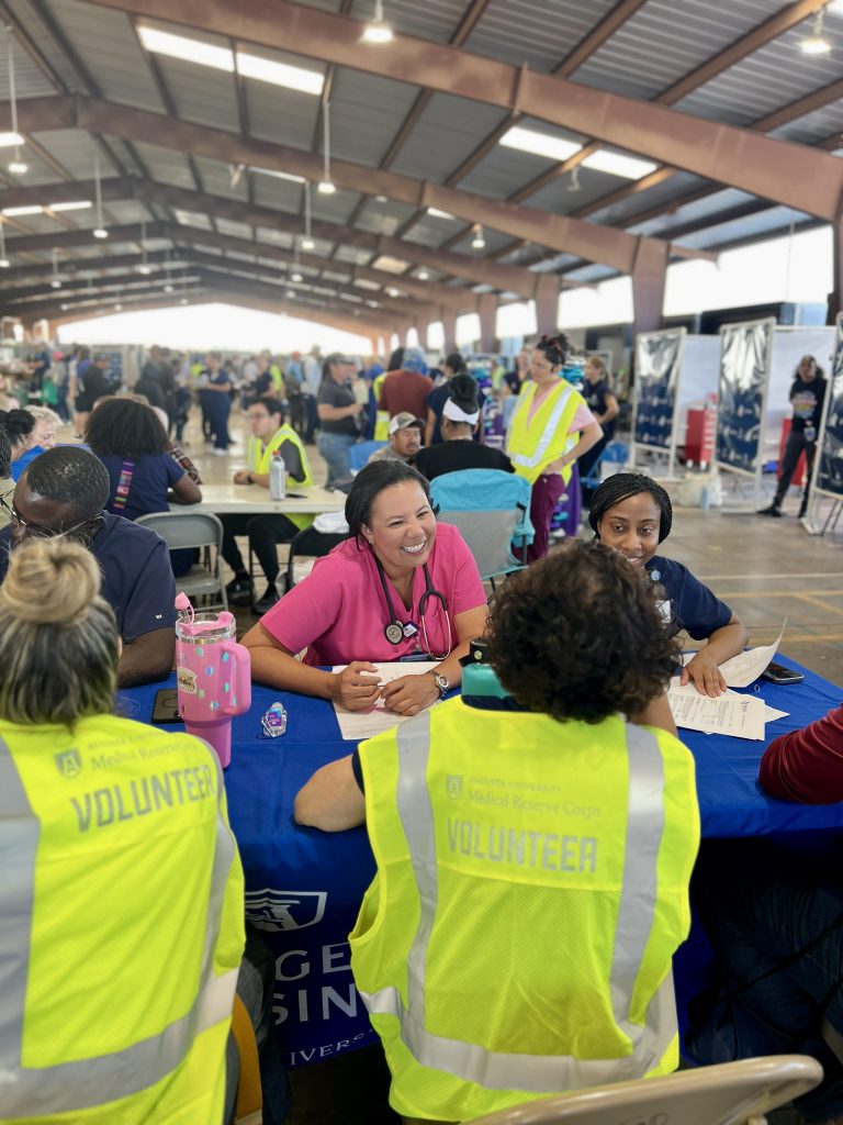A woman in scrubs sits at a table. She is smiling warmly at one of the volunteer interpreters as they speak with a participant who is just off camera.