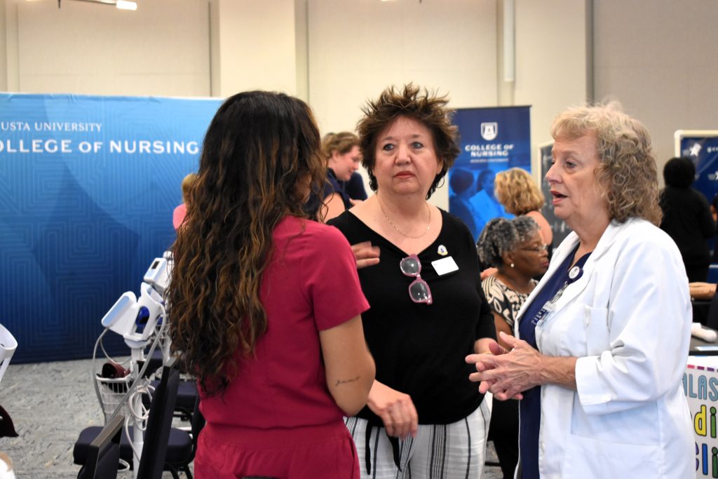 Three women talk to each other, a health fair and banners are in the background.