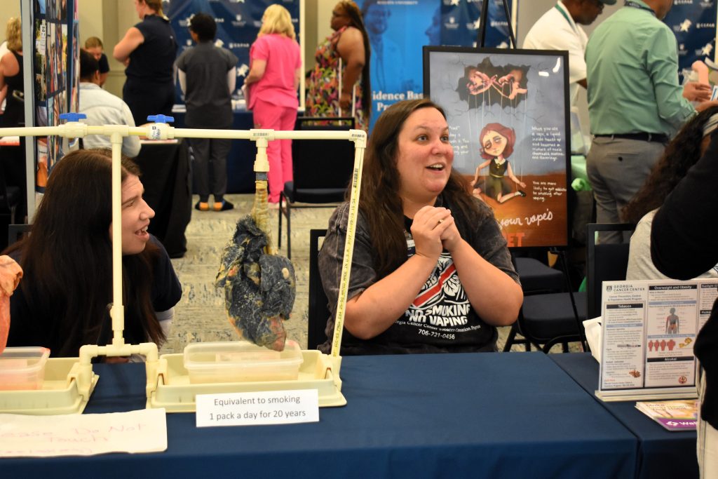 Two women sit behind a table. One of them is speaking and is seated next to a model of an unhealthy lung that has smoked 1 pack a day for 20 years.