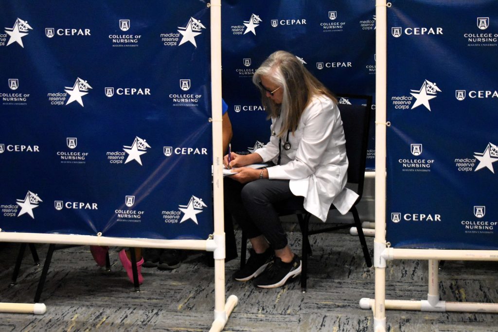 A woman in a white coat sits in a chair, she is speaking to a child who is blocked by privacy screens.
