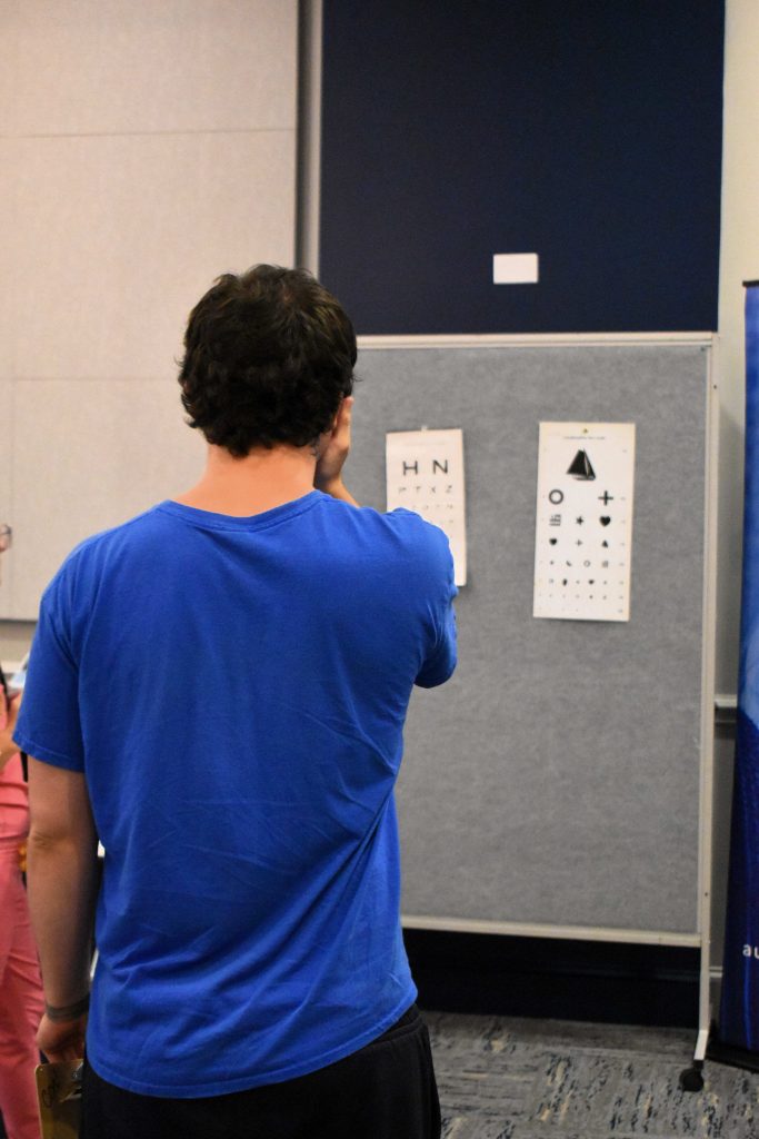 A young man, photographed from the back, covers an eye and performs a vision test. Across from him is a board with print outs of letters and symbols.