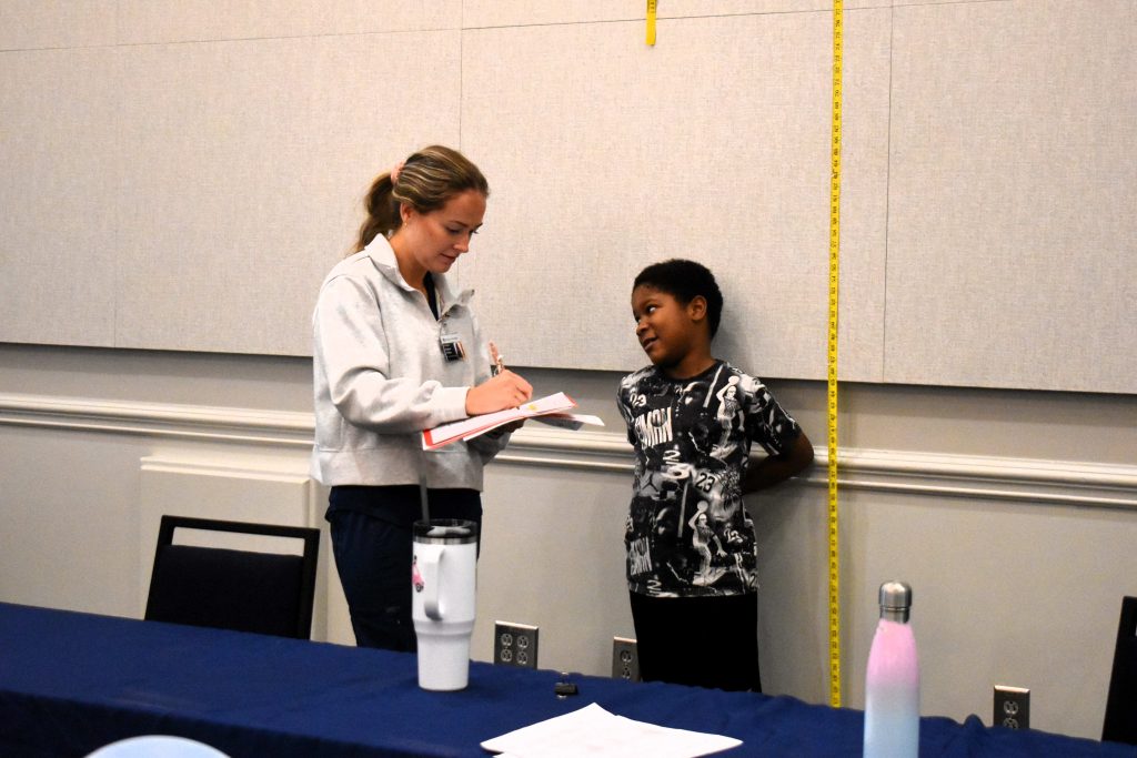 A young child speaks to a healthcare provider. He is standing next to a measuring tape, getting his height measured.