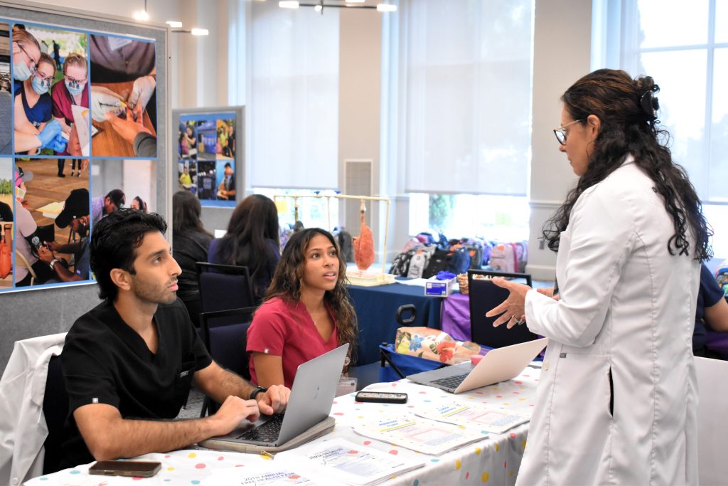 Two people sit at an information table. They are talking to a woman in a white coat.