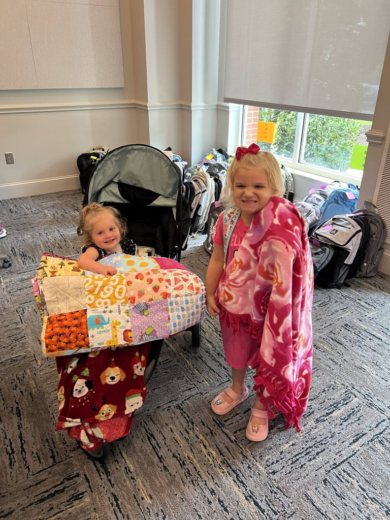 Two young girls, one in a stroller, hold up their blankets and smile for the camera.