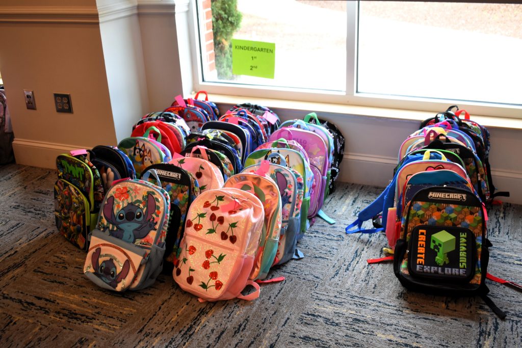 Three rows of children's backpacks sit on the ground next to a window