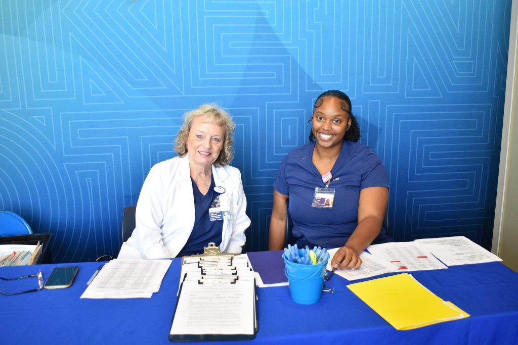 Two women sit behind a table. There is a step-and-repeat behind them, and they are smiling at the camera.