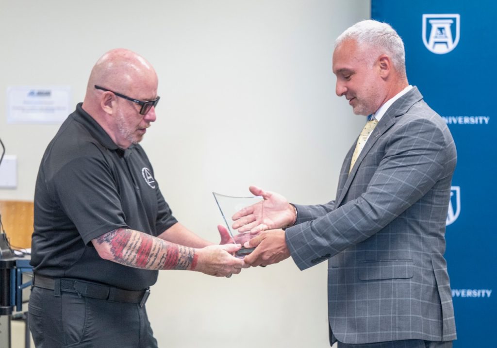 Two men stand together, with one handing the other an award. 
