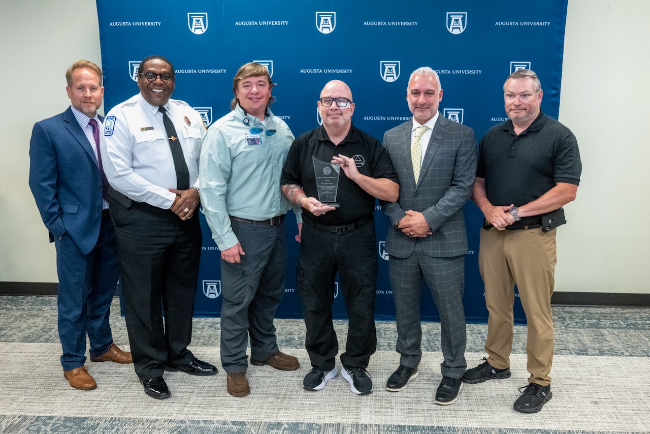 six men stand together smiling with one holding an award