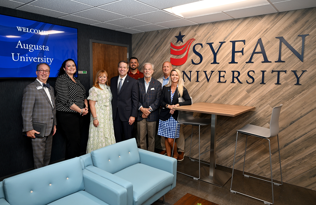 Men and women stand behind a sofa and in front of a monitor and large letters