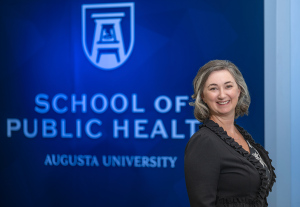 A professionally dressed woman stands in front of a wall that reads "School of Public Health."