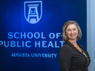 A professionally dressed woman stands in front of a wall that reads "School of Public Health."