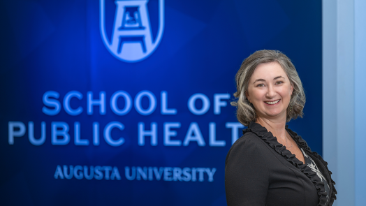 A professionally dressed woman stands in front of a wall that reads "School of Public Health."