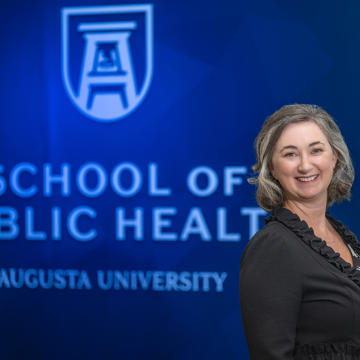A professionally dressed woman stands in front of a wall that reads "School of Public Health."