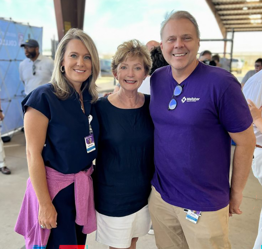 Three people pose together, smiling at the camera. They are outside surrounded by a crowd of people.