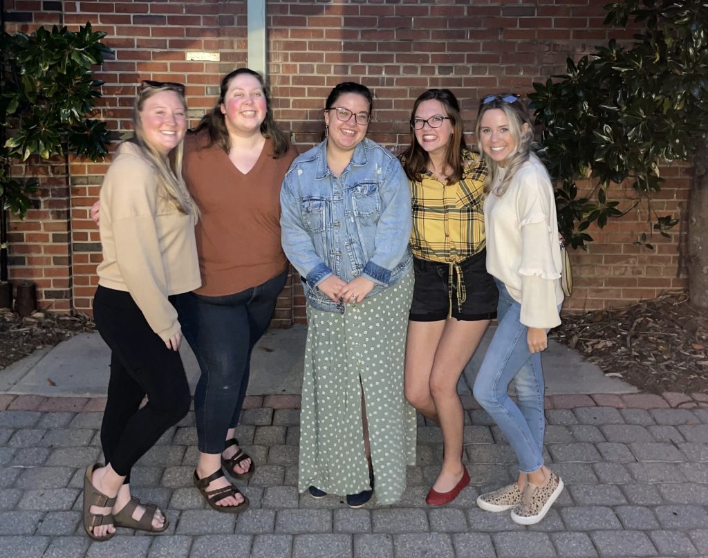 All five women pose together, smiling at the camera.