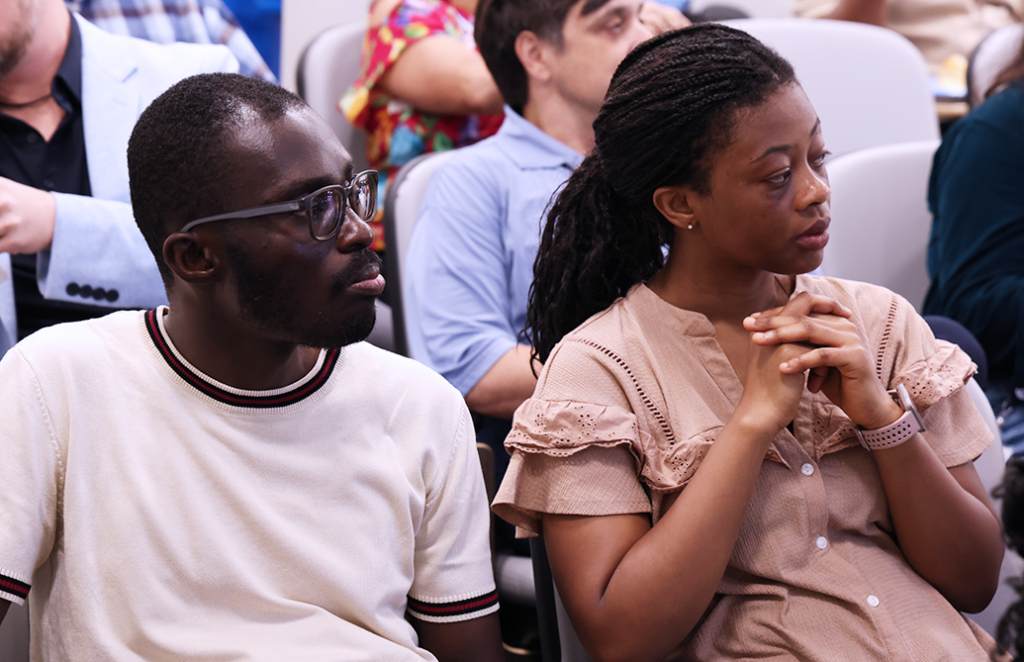 A man and a woman sit beside each other in a row in an auditorium and listen to a speaker.