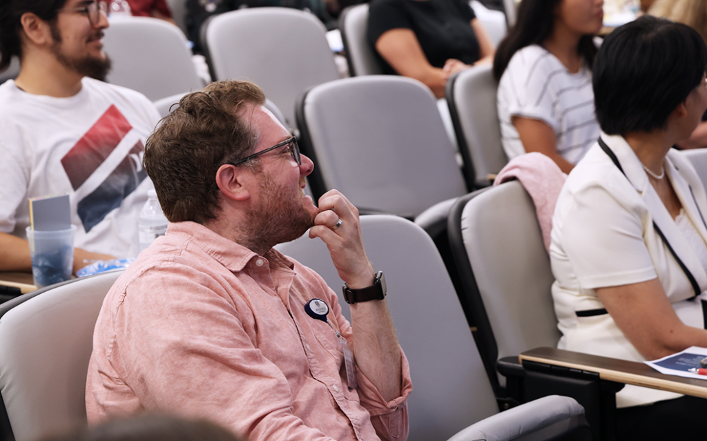 A man sits in a chair in an auditorium and smiles.