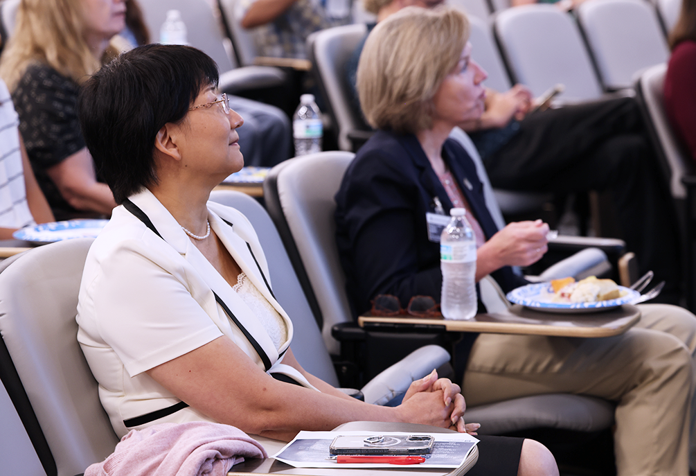 Two women sit in chairs in a row in an auditorium and listen to a speaker.
