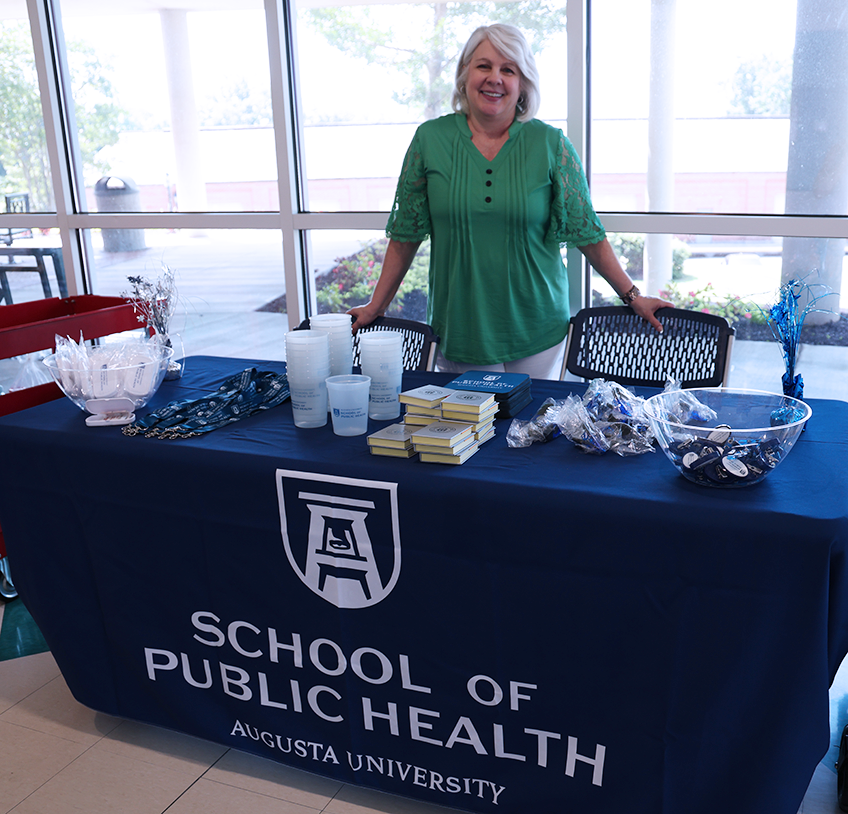 A woman stands at a table with a cloth that reads "Augusta University School of Public Health" and smiles.