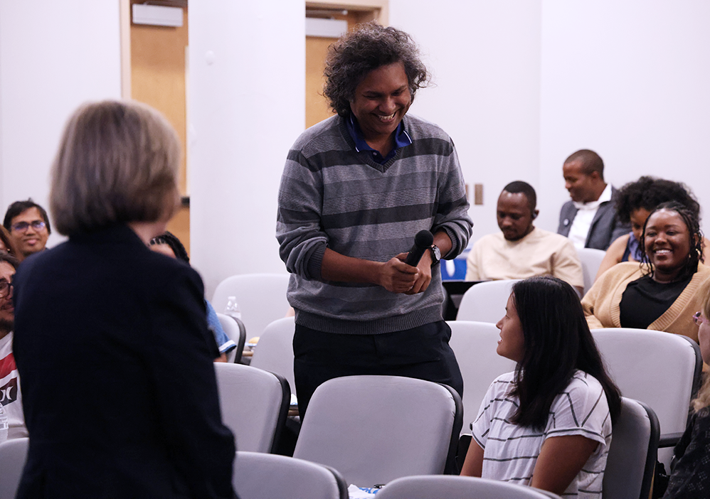 A man holding a microphone stands in a row of people in an auditorium and laughs.