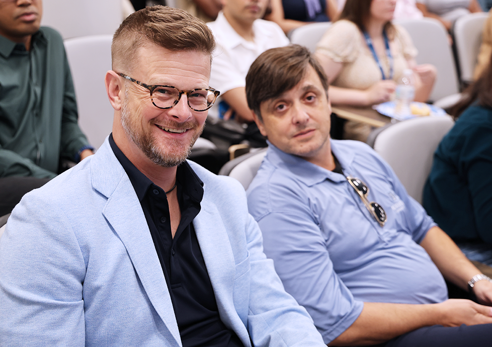 Two men sitting in seats in an auditorium smile at the camera.