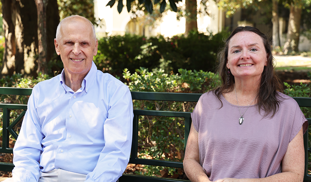 A man and a woman sit on a park bench and smile at the camera.