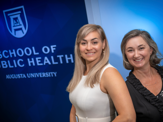 Two professionally dressed woman standing in front of a wall that reads "School of Public Health" smile at the camera.