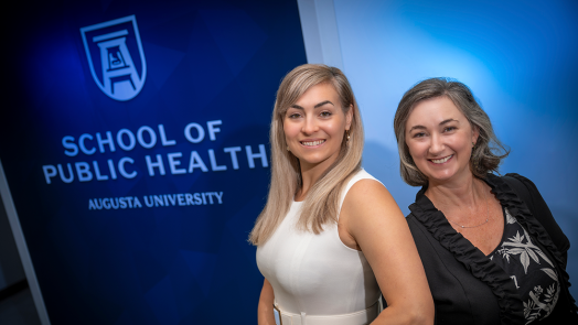 Two professionally dressed woman standing in front of a wall that reads "School of Public Health" smile at the camera.
