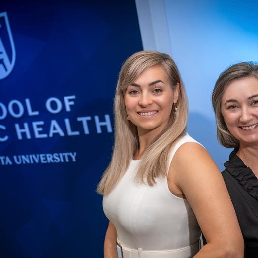 Two professionally dressed woman standing in front of a wall that reads "School of Public Health" smile at the camera.