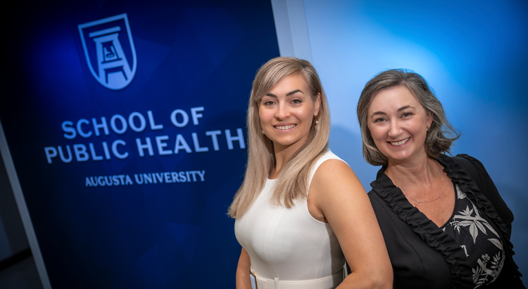 Two professionally dressed woman standing in front of a wall that reads "School of Public Health" smile at the camera.