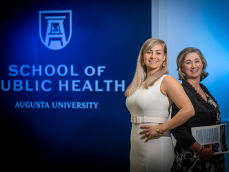 Two professionally dressed woman standing in front of a wall that reads "School of Public Health" smile at the camera.