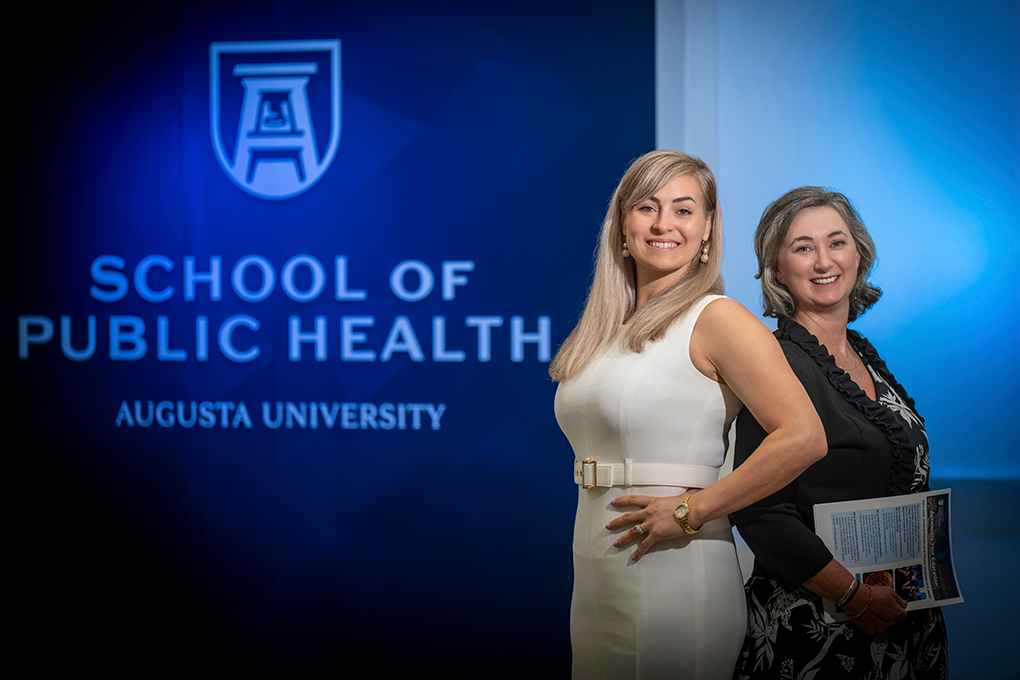 Two professionally dressed woman standing in front of a wall that reads "School of Public Health" smile at the camera.