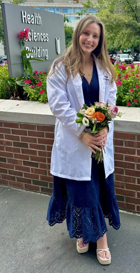 Young woman stands in front of Health Sciences Building sign, smiling and holding a boquet of flowers.