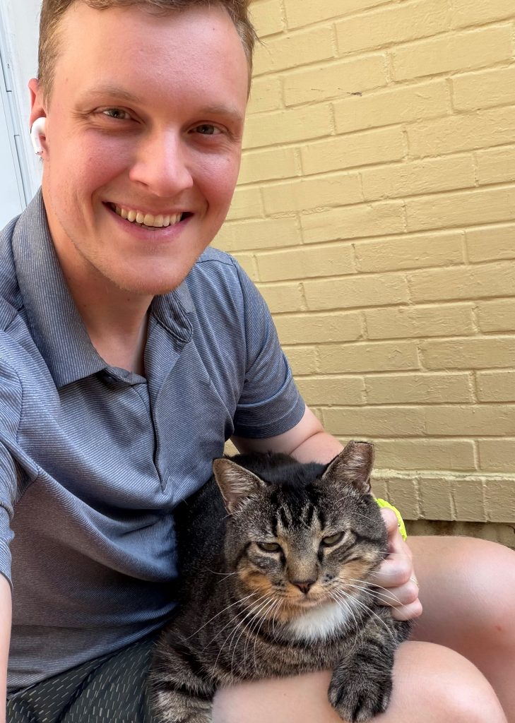 A man in a polo sits outside near a brick wall, posing with a cat in his lap